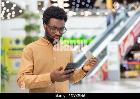 Junger erwachsener Mann in Brille, der ein Smartphone und eine Kreditkarte hält, beim Online-Banking oder beim digitalen Kauf in einem modernen Einkaufszentrum besorgt aussieht Stockfoto