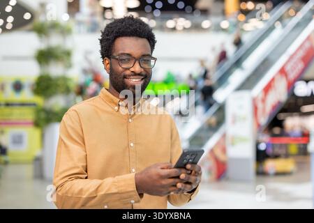 Junger Mann, der ein Smartphone benutzt und lächelt, in einem modernen Einkaufszentrum steht, sich mit Technologie verbindet und einen urbanen Lebensstil erlebt Stockfoto