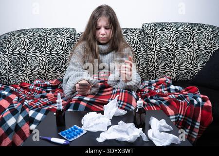Das kranke Mädchen, in eine Decke gewickelt, sitzt auf einem Sofa und hält ein Glas Wasser und Pillen. Tisch mit Nasenspray, Thermometer und Servietten im Vordergrund Stockfoto