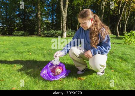 Ein Mädchen, das Schutzhandschuhe trägt, hockt auf grünem Gras und nimmt Müll in einen violetten Beutel. Sie hilft, im Herbst einen sonnigen öffentlichen Park zu säubern Stockfoto