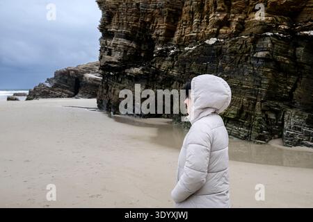 Frau, die auf dem nassen Sand steht, die geschichteten Felsformationen und Klippen von Cathedrals Beach beobachtet und die dramatische Naturlandschaft während des Sieges genießt Stockfoto