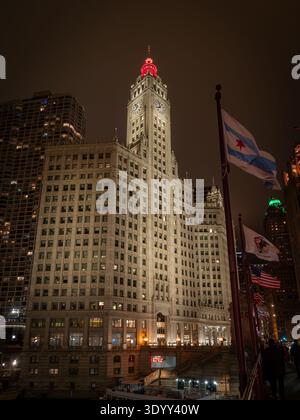 Das Wrigley Building in der Innenstadt von Chicago ist nachts beleuchtet mit roter Turmbeleuchtung und schwenkenden Chicagoer Flaggen Stockfoto