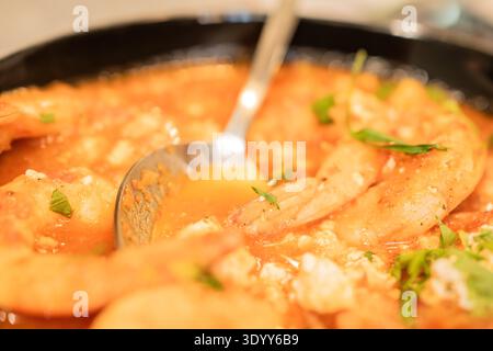 Eine Schüssel mit Garnelen, die in einer Sauce gekocht sind, steht auf einem Tisch. Das Gericht enthält Kräuter und Zutaten, die mit den Garnelen gemischt werden. Die Umgebung ist bereit für das Abendessen. Stockfoto