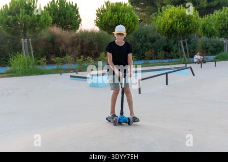 Selbstbewusster Jugendlicher in schwarzem Hemd und Mütze posiert auf blauem Roller im Skatepark zur goldenen Stunde. Konzept der kühlen städtischen Jugend, Unabhängigkeit und aktiv Stockfoto