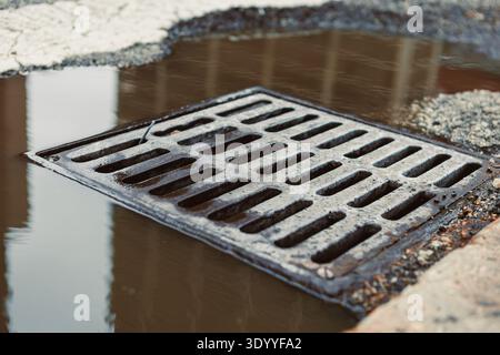 Wasser sammelt sich in einer Pfütze in der Nähe einer Metallabflussabdeckung auf einer Stadtstraße, nachdem es tagsüber regnet. Stockfoto