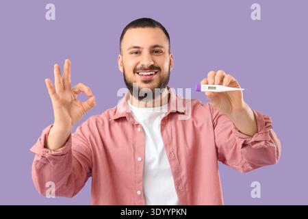 Hübscher junger Mann mit Thermometer, das OK-Geste auf Flieder-Hintergrund zeigt Stockfoto