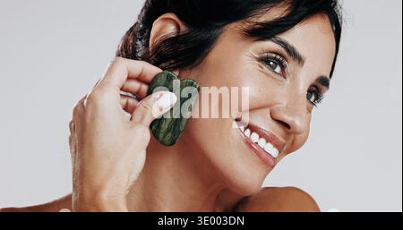 Frau, Gua Sha und Lächeln mit Schönheit im Studio für Zirkulation, Denken und Hautpflege auf weißem Hintergrund. Reifer Mensch, Steinwerkzeuge und Kosmetik Stockfoto