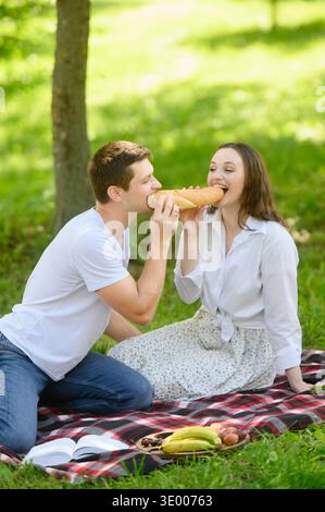 Ein Paar genießt ein Picknick auf einer Decke unter einem Baum, ein entspanntes Paar genießt einen Imbiss im Freien. Liebende teilen Brot, beißen gleichzeitig hinein und lachen Stockfoto