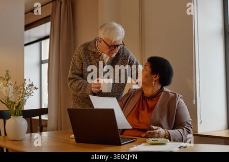 Senior-kaukasischer Mann, der mit Senior Black Frau spricht, die zu Hause an Laptop arbeitet Stockfoto