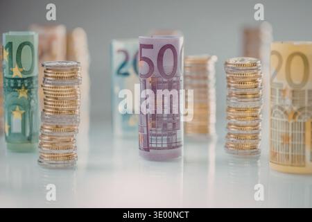 Gerollte Euro-Banknoten in verschiedenen Stückelungen stehen neben Stapeln von Euro-Münzen auf einer glänzend weißen Oberfläche und spiegeln sich ordentlich wider. Stockfoto