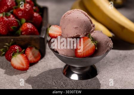 Ein Erdbeersorbet aus drei Messlöffeln, serviert in einer kleinen Schüssel aus Metall, garniert mit halbierten frischen Erdbeeren. Im verschwommenen Hintergrund, eine Schachtel reifer berri Stockfoto