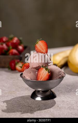 Ein Erdbeersorbet aus drei Messlöffeln, serviert in einer kleinen Schüssel aus Metall, garniert mit halbierten frischen Erdbeeren. Im verschwommenen Hintergrund, eine Schachtel reifer berri Stockfoto