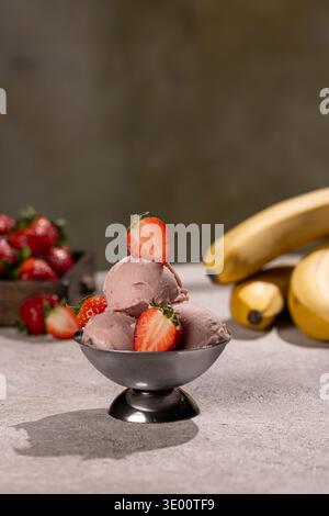 Ein Erdbeersorbet aus drei Messlöffeln, serviert in einer kleinen Schüssel aus Metall, garniert mit halbierten frischen Erdbeeren. Im verschwommenen Hintergrund, eine Schachtel reifer berri Stockfoto