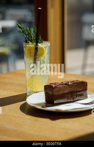 Erfrischende Limonade mit Rosmarin und Zitronenscheibe im Glas auf dem Kaffeetisch. Dessert mit Schokoladenkuchen im Sonnenlicht, kaltes Getränk, Café-Lifestyle-Konzept Stockfoto