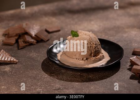 Nahaufnahme einer großen Schaufel cremiges Schokoladeneis, bestreut mit feinen Schokoladenspänen, auf einer mattschwarzen Platte. Das gestylte Studio wurde im Krieg gedreht Stockfoto