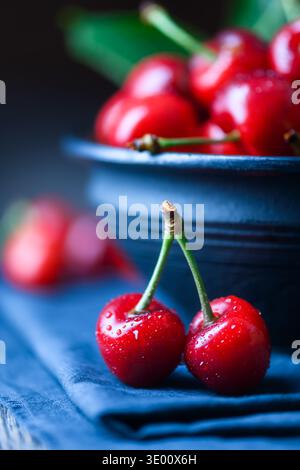 Paar reife Kirschen auf blauer Serviette mit Keramikschale im Hintergrund. Frische, glänzende rote Früchte, geringe Tiefe, Sommerkonzept Stockfoto
