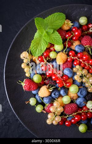 Gemischte Sommerbeeren auf dunklem Teller mit Minzblättern, Blick von oben. Heidelbeeren, Himbeeren, Johannisbeeren, Stachelbeeren und Maulbeeren, Bio-Sortiment der Saison Stockfoto