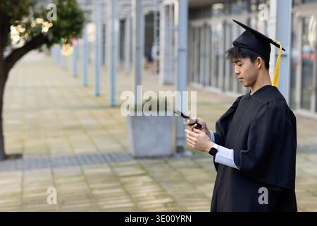 Asiatischer Mann, der auf der plaza steht und ein Smartphone hält, trägt ein Kleid, eine Mörtelplatte mit Quaste, einen Kopierraum. Absolvent, Mütze, Bademantel, Telefon, Uhr, Campus, im Freien Stockfoto