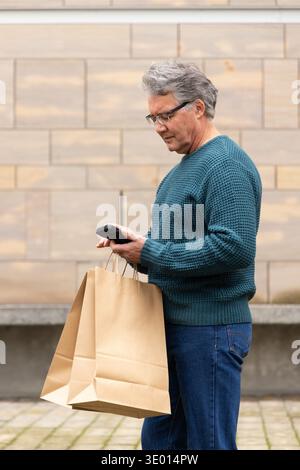 Älterer Mann mit blauem Pullover und Brille, der braune Papiertüten mit Telefon an gekachelten Wänden hält Stockfoto