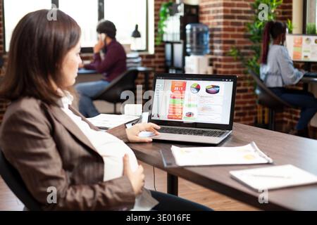 Kaukasische schwangere Frau mit Hand auf dem Magen prüft die Marktanalyse des Unternehmens auf dem Laptop. Werdende Dame, die digitale Geräte am Bürotisch verwendet und Finanzstrategien im Startup-Büro recherchiert. Stockfoto
