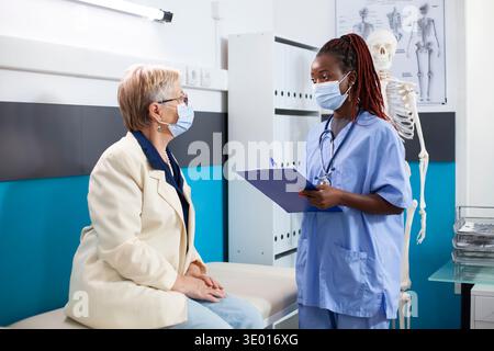 Afroamerikanische Krankenschwester mit Klemmbrett steht am Krankenhausbett und diskutiert mit älteren weißen Frauen, wie beide Gesichtsmasken tragen. Pensionierter Patient hört während eines Arzttermins aufmerksam auf junge Ärzte zu. Stockfoto