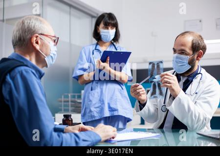 Arzt erklärt Röntgenergebnisse älteren Männern, während weibliche medizinische Assistentin Notizen während des Arzttermins macht. Krankenschwester mit Klemmbrett neben dem Arzt, der dem Rentner eine Röntgenaufnahme zeigt. Stockfoto