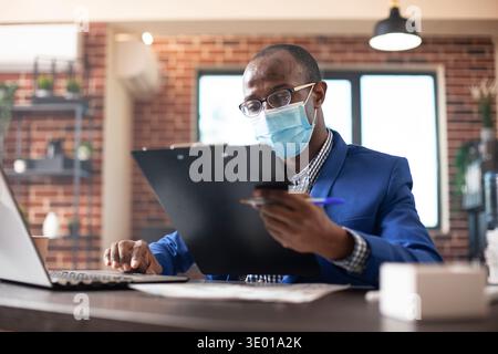 Großaufnahme des Unternehmers in der Gesichtsmaske, das Clipboard hält und Projektdokumente am Schreibtisch überprüft, Präsentationen mit Laptop planen. afroamerikaner liest und bereitet Firmenpapiere vor. Stockfoto