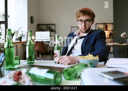 Porträt eines jungen kaukasischen Mannes, der am Schreibtisch sitzt, umgeben von Bierflaschen Stockfoto