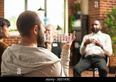 Nahaufnahme eines bärtigen Mannes, der während der Gruppentherapie mit den Fingern unterschiedliche Zeichen macht. Junger weißer Kerl, der im Kreis sitzt und Handgesten im Mauerraum macht, während die Leute im Hintergrund diskutieren. Stockfoto