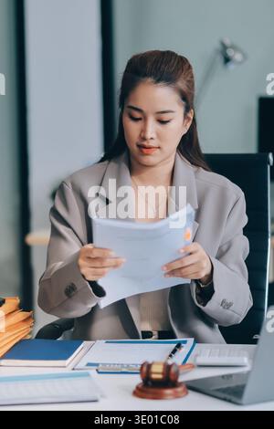 Selbstbewusste Anwältin bei Desk: Eine junge asiatische Anwältin arbeitet selbstbewusst an ihrem Laptop, umgeben von juristischen Symbolen. Sie strahlt Professionalität aus Stockfoto