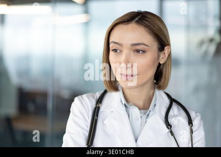 Die Ärztin blickt weg, denkt über medizinische Aufgaben nach, trägt einen weißen Reverskmantel und ein Stethoskop über den Schultern, was für Gesundheit, Medizin und Wellness steht Stockfoto