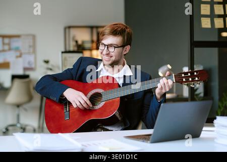 Kaukasischer junger Erwachsener, der Akustikgitarre spielt, während er lächelnd am Schreibtisch sitzt Stockfoto