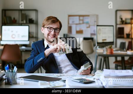 Junger Kaukasier lächelt, während er einen Stapel Dollar-Scheine im Amt hält Stockfoto