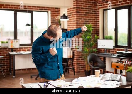 Fröhlicher junger Unternehmer steht neben Tisch und hebt seine Fäuste vor Freude in einem gemauerten Wandzimmer. Bärtiger Mann feiert erfolgreiche Geschäftspräsentation und Projektsieg im Startup-Büro. Stockfoto