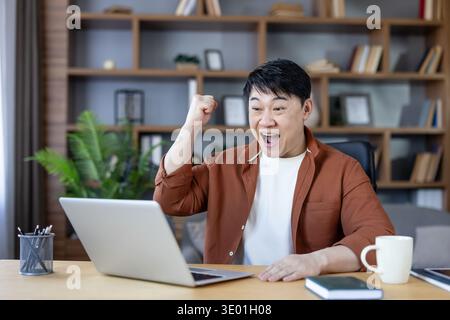 Asiatischer Mann, der anfeuert und seine Faust zum Feiern hebt, während er seinen Laptop ansieht und Erfolg, Sieg und Glück erlebt, indem er von seinem Home Office aus aus arbeitet Stockfoto