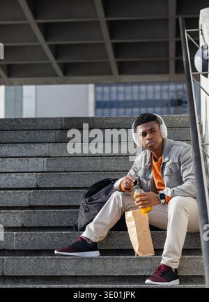 Afroamerikaner sitzt auf Betontreppen am plaza und hält Orangengetränk mit Tasche, Kopierraum. Urban, Treppenhaus, Rucksack, Kopfhörer, Smartwatch Stockfoto