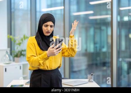 Muslimische Geschäftsfrau im Hijab in einem modernen Büro, die an ihrem Schreibtisch steht und mit Schock und Frustration auf unerwartete Nachrichten auf ihrem Smartphone reagiert, gestresst und besorgt Stockfoto