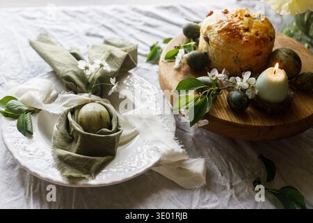 Frohe Ostern! Stilvolle Eier in Hasenohren Serviette auf Teller, osterkuchen auf dem Ständer, Frühlingsblumen und Kerze. Osterfeierungstafel, Feiertage Stockfoto