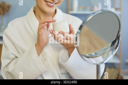 Lächelnde Frau in weißem Gewand, die ein Glas Creme hält und Kosmetik in der Nähe des Spiegels ausprobiert, um Hautpflege zu machen Stockfoto