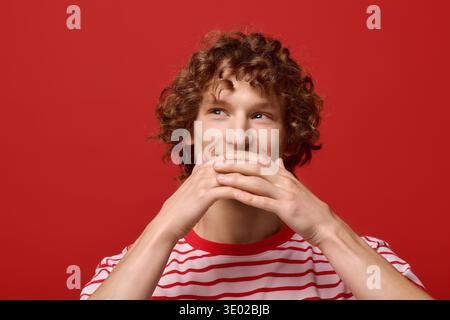 Ein stilvoller junger Mann mit lockigen Haaren bedeckt seinen Mund und lächelt in einem schicken roten Studio-Hintergrund. Stockfoto