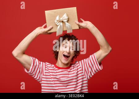 Ein junger Mann mit lockigen Haaren hält freudig ein Geschenk über dem Kopf in einem hellroten Studio-Setting. Stockfoto