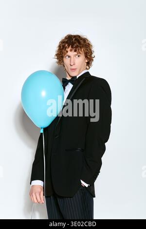 Ein glücklicher junger Mann mit lockigen Haaren hält einen blauen Ballon in einem schicken Smoking. Stockfoto
