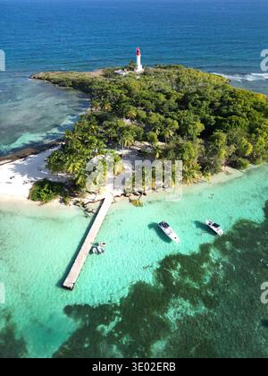 Blick aus der Vogelperspektive auf einen Leuchtturm, der in üppigem Grün auf einer tropischen Insel steht, umgeben von türkisfarbenem Wasser und Booten, Guadeloupe, Guadeloupe. Stockfoto