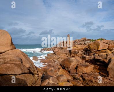 Der Leuchtturm Mean Ruz erhebt sich über massiven Granitblöcken in Tregastel an der Pink Granite Coast in der Bretagne, Frankreich, mit Blick auf Wellen und die ferne isla Stockfoto