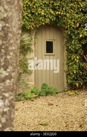 Bogenförmige Holztür mit Fenster in Steinmauer, üppiger Efefeuüberzug, Schotterweg, der zu einem geheimen Garteneingang führt. Cotswolds, England, Vereinigtes Königreich Stockfoto
