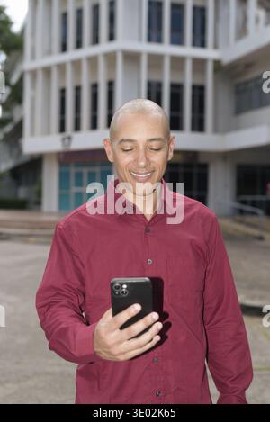 Glatzkopf italienischer Mann in burgunderrotem Hemd, der sein Smartphone überprüft, während er vor dem Universitätsgebäude bei hellem Sonnenlicht steht. Das Konzept des digitalen Kombi Stockfoto