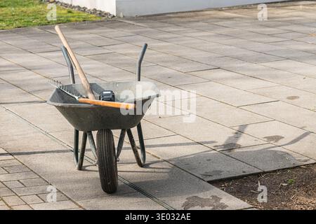 Eine Schubkarre steht auf einem Steinweg im Hof. In der Schubkarre befindet sich ein Rechen. Das Gras ist grün und die Sonne wirft Schatten auf den Grou Stockfoto