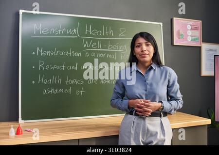 Porträt einer jungen erwachsenen hispanischen Frau, die im Klassenzimmer vor der Tafel mit Informationen über die Menstruation Gesundheit und das Wohlbefinden steht, Hände zusammenhält und in die Kamera blickt Stockfoto