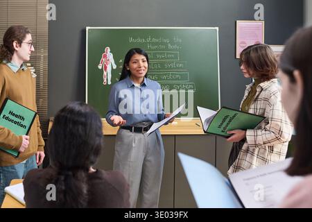 Junge Erwachsene hispanische Frau, die vor der Tafel steht und der Gruppe junger erwachsener Studenten die Systeme des menschlichen Körpers erklärt, die während des Unterrichts Gesundheitsaufklärungsmappen halten Stockfoto