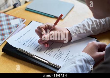 Teenager, der mit Stift auf gedrucktem Prüfungspapier Antworten schreibt, mit Klemmbrett und Notizbüchern am Schreibtisch sitzt und fokussierte Handbewegungen und Teilgesicht in Bildungseinrichtungen zeigt Stockfoto